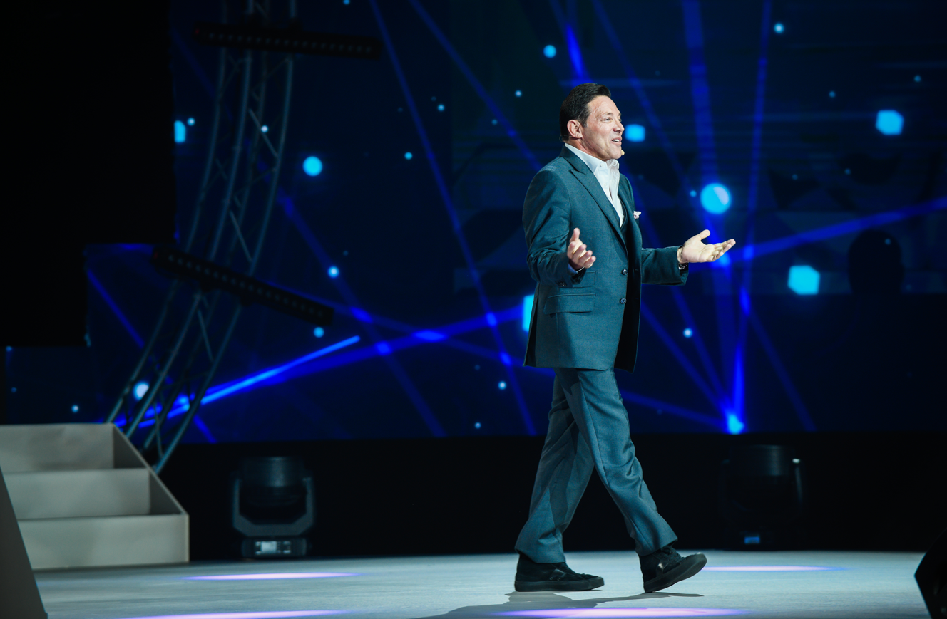 Jordan Belfort speaking to an audience on a blue-lit stage at a previous event.